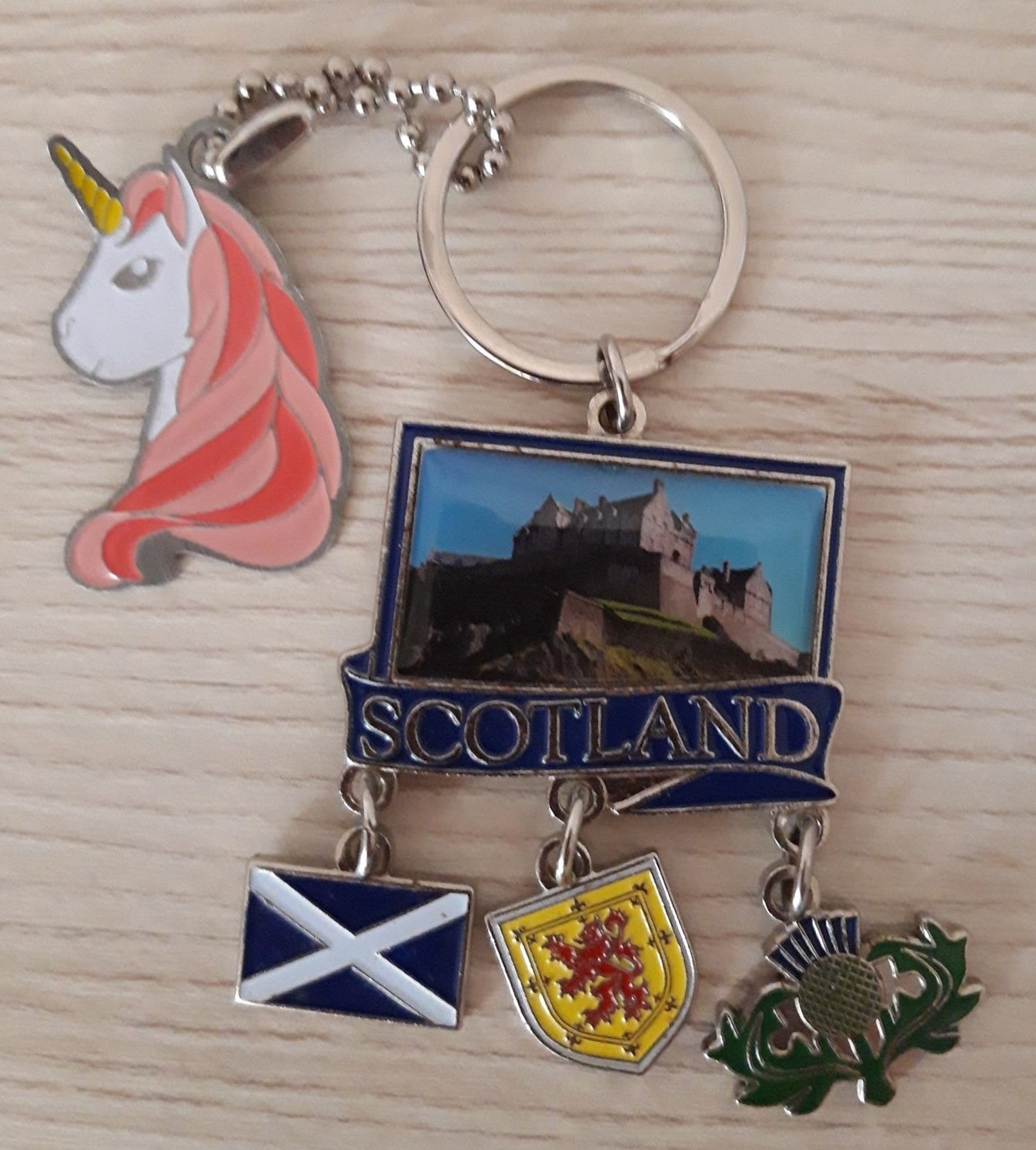 A metal unicorn head key ring with long floating hair (light pink and dark pink). She is carrying a metal Scotland key ring of Edinburgh Castle with the words Scotland across it, which is holding three metal trinkets: the Scotland flag, lion rampant and a Scottish thistle