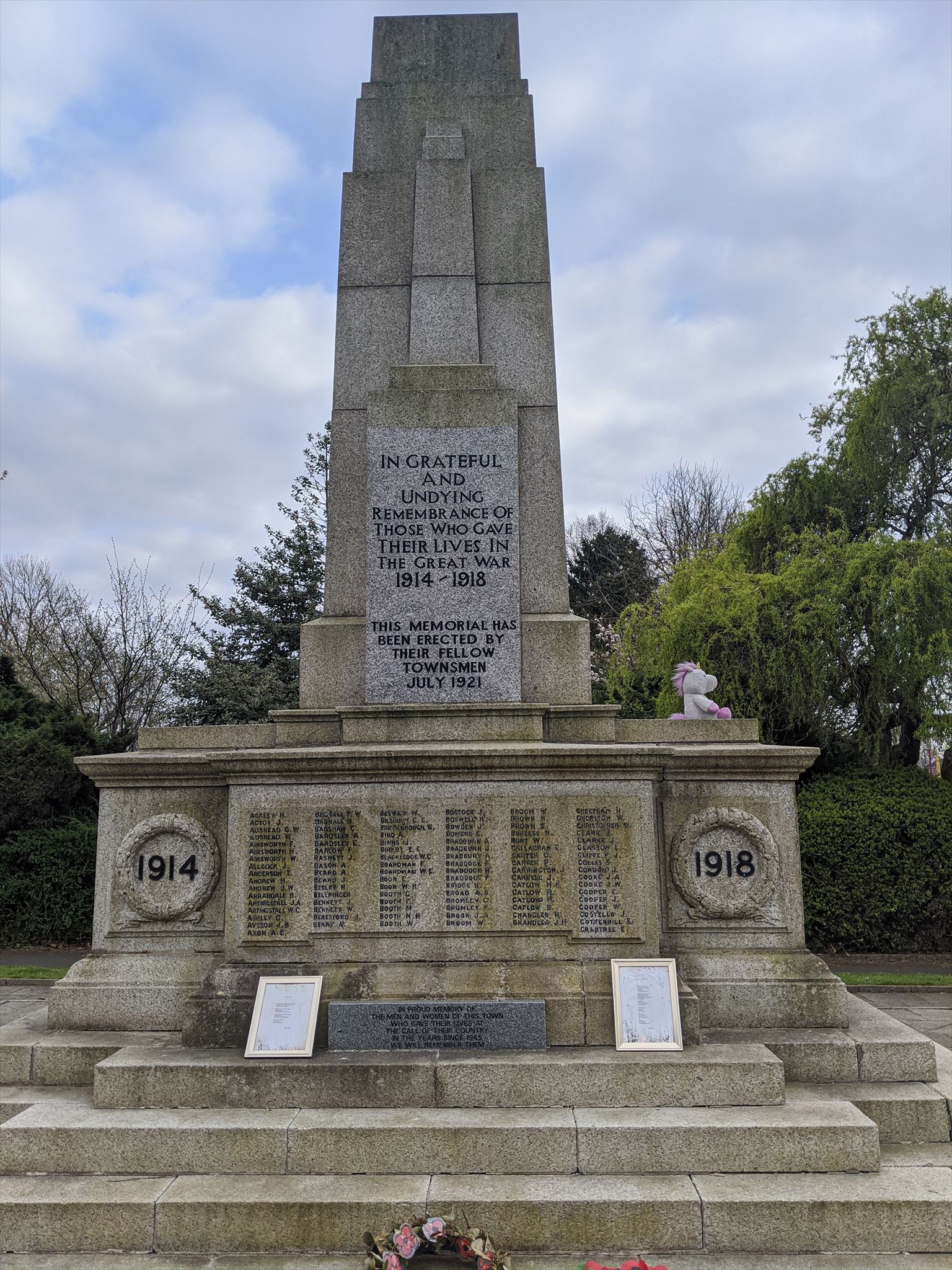 Photograph of Victoria Park War Memorial. Two inscriptions are clearly visible (see below). On the front face of the monument, a list of over 100 names split into 6 columns. On the left and right of the memorial are two dates, 1914 and 1918. Sitting on the memorial is a small stuffed toy left in tribute. Flower wreaths are visible at the base of the steps of the monument.