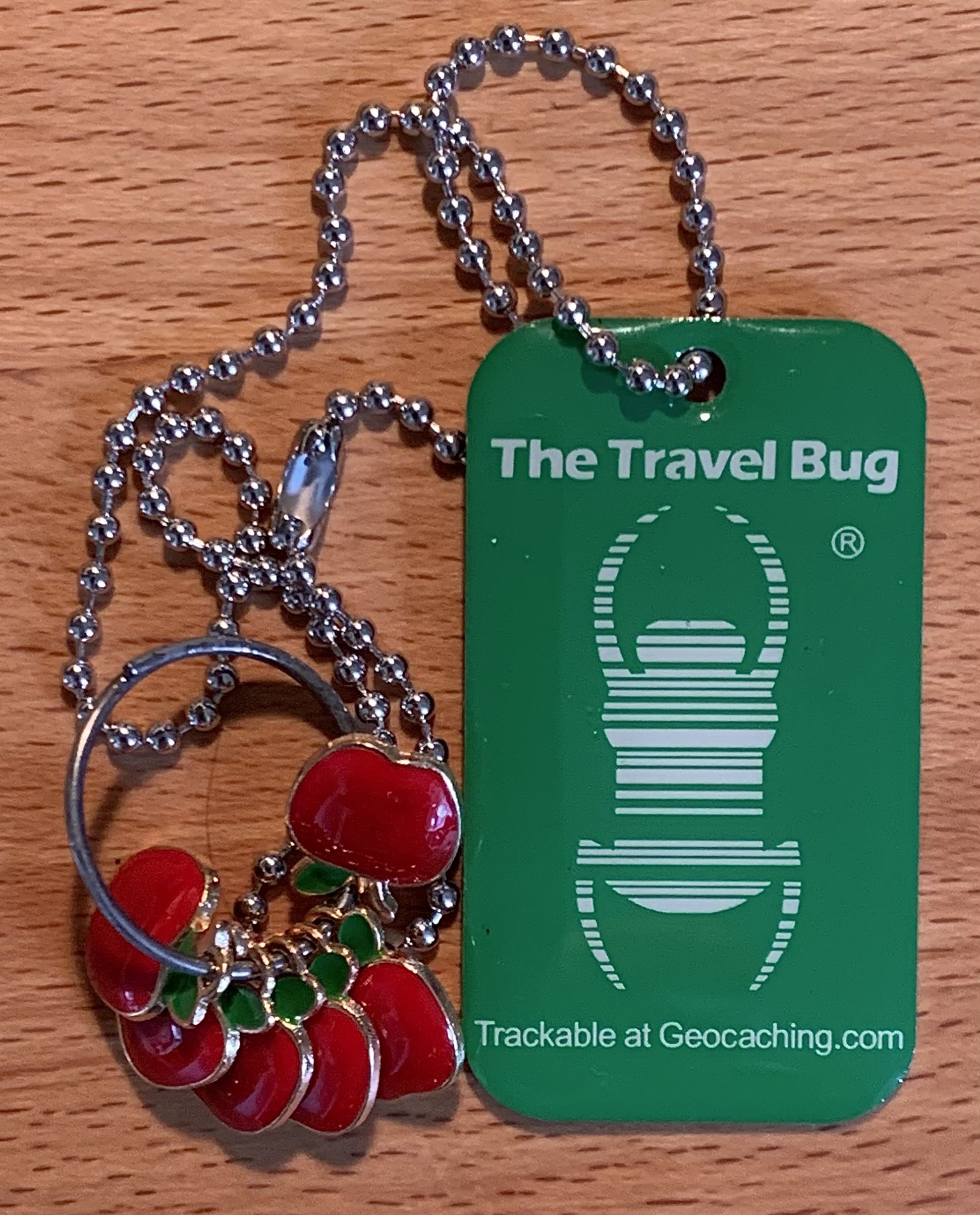 6 rote Äpfelchen aus Metall an einem Schlüsselring, die an der Kette eines grünen, rechteckigen Travelbugs hängen