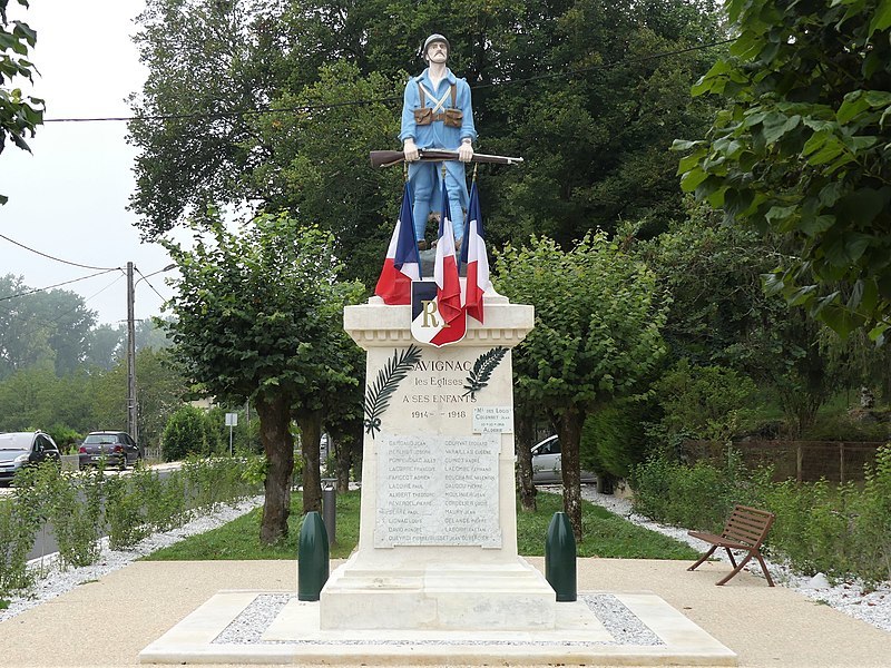 Photo montrant la statue du Soldat de Pourquet. Un homme en tenue bleue de soldat se tient debout, les jambes écartées. Il porte un casque de soldat en métal. Dans ses mains, un fusil. A ses pieds, des drapeaux Français.