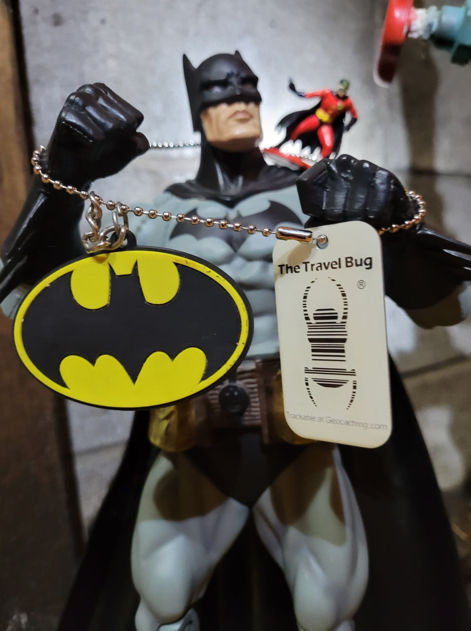 This TRACKABLE is a Glow-in-the-Dark Travel Bug.  It glows a greenish color in the dark after being exposed to light.  I attached a small one and a half inch Robin figure, Batman’s side-kick.  Robin has a standing pad connected to his feet.  I threaded the chain from the Trackable Travel Bug between Robin’s legs to attach him to the trackable.   I also attached the Trackable's chain to a Bat Emblem Keychain that is rubber, and has a short chain on top.  This Bat Emblem is an oval about two inch tall and about three inches wide. The rubber emblem is about one eighth inch thick.  The Bat Emblem has a Yellow background with a Black Bat filling the center to the edges.   My son has a Batman posable figure that is about eleven inches tall.  I posed the Robin figure on Batman’s Left shoulder, with the chain of the trackable around Batman’s neck.  Batman is posed with his two fists up in a fighting position.  He has the chain of the Trackable around both fists, with the Bat Emblem hanging from his Right fist, and the Tracking Bug hanging from his Left fist.   I took their photo in our basement, with a cinder block wall in the background, next to pipes and a red valve.  I shined a flashlight beam behind Batman on the wall, so that Batman and the three parts of the Trackable would appear illuminated in the foreground with the light behind them.  I took the photo from below Batman’s waist, looking up so he would look big.  This effect was also to simulate Batman and the ensemble attached to the Trackable as if they were in a back alley or chemical plant with dramatic lighting.   