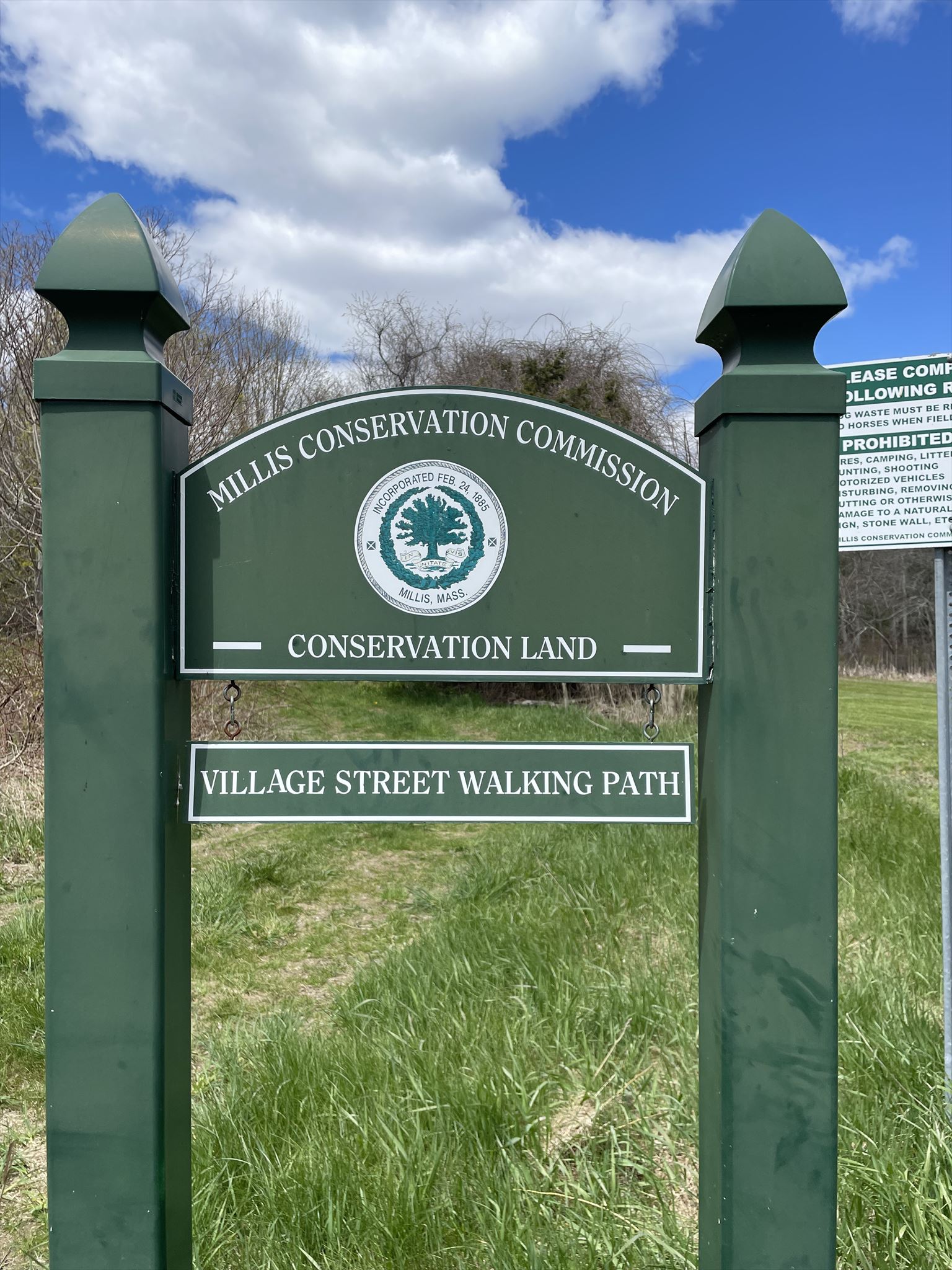 photo of trailhead signage - Millis conservation commission, village street walking path. Printed on green wooden sign. Sign is in a grassy field with trees and a blue cloudy sky in the background.